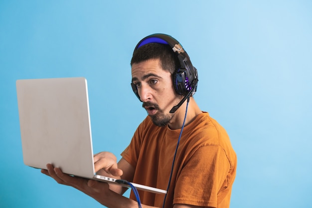 Jeune bel homme avec jeu de casque gamer isolé sur fond bleu à l'aide d'un ordinateur portable