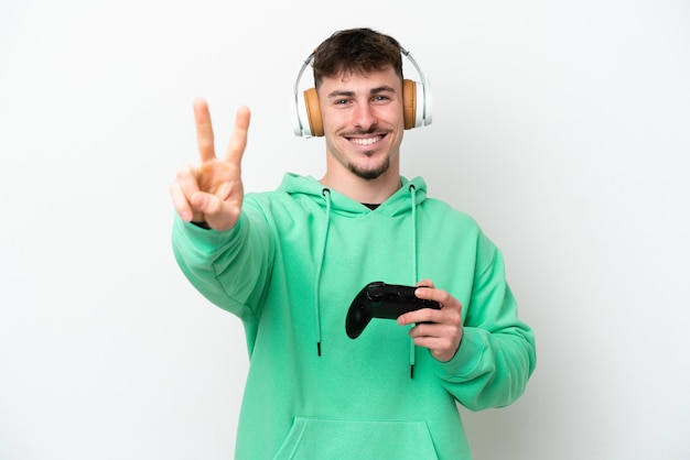 Jeune bel homme jouant avec un contrôleur de jeu vidéo isolé sur fond blanc souriant et montrant le signe de la victoire