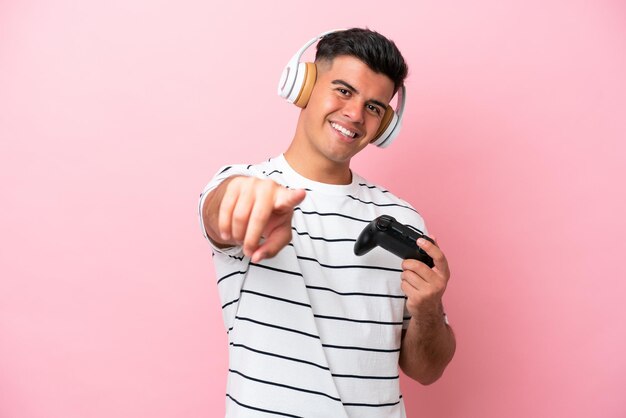 Jeune bel homme jouant avec un contrôleur de jeu vidéo isolé sur fond rose pointant vers l'avant avec une expression heureuse