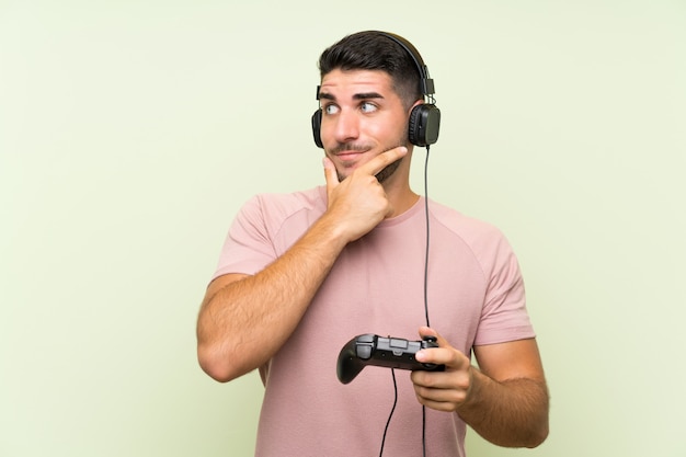 Jeune bel homme jouant avec un contrôleur de jeu vidéo sur un mur vert isolé, pensant une idée