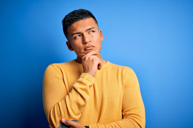 Jeune bel homme latin portant un pull décontracté jaune sur fond bleu isolé avec la main sur le menton pensant à la question expression pensive Souriant avec un visage pensif Concept de doute