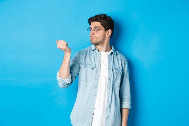 Jeune bel homme à lunettes regardant ses ongles, vérifiant la manucure, debout sur fond bleu