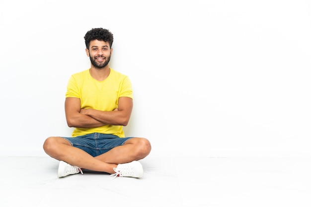 Jeune bel homme marocain assis sur le sol sur fond isolé avec les bras croisés et heureux