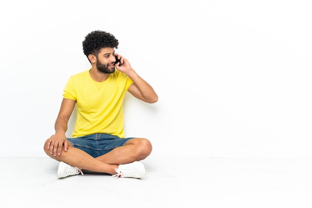 Jeune bel homme marocain assis sur le sol sur fond isolé en gardant une conversation avec le téléphone mobile avec quelqu'un