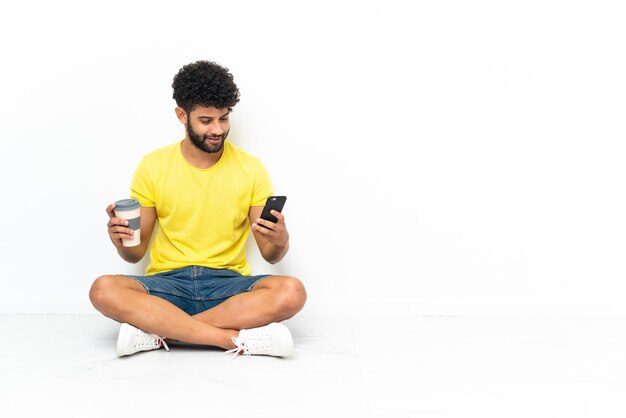 Jeune bel homme marocain assis sur le sol sur un mur isolé tenant du café à emporter et un mobile