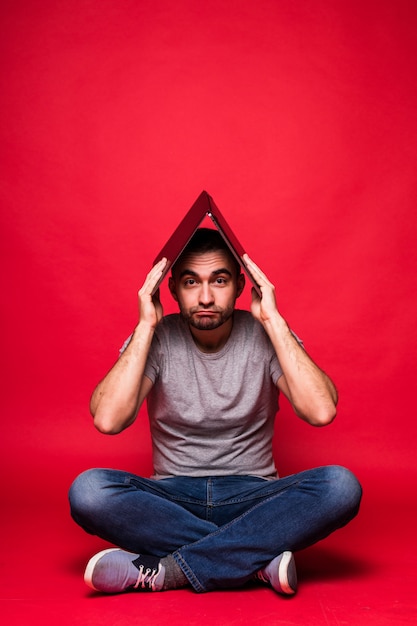 Jeune bel homme avec ordinateur portable sur la tête assis sur le sol isolé sur fond rouge