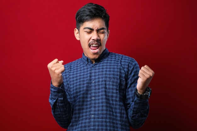 Jeune bel homme portant une chemise décontractée debout sur un fond rouge isolé très heureux et excité faisant le geste du gagnant avec les bras levés, souriant et criant pour le succès. Notion de célébration