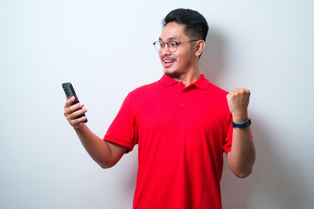 Jeune bel homme portant une chemise décontractée et des lunettes debout très heureux et excité faisant le geste du gagnant avec les bras levés en souriant tout en regardant son téléphone portable