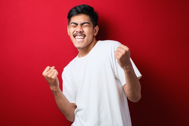 Jeune Bel Homme Portant Un T-shirt Blanc Debout Sur Fond Rouge Isolé Très Heureux Et Excité Faisant Le Geste Du Vainqueur Avec Les Bras Levés, Souriant Et Criant Pour Le Succès. Notion De Célébration