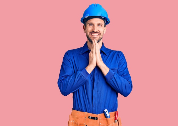 Jeune bel homme portant un uniforme de travailleur et un casque priant avec les mains jointes demandant pardon souriant confiant