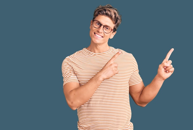 Jeune bel homme portant des vêtements décontractés et des lunettes souriant et regardant la caméra pointant avec les deux mains et les doigts sur le côté