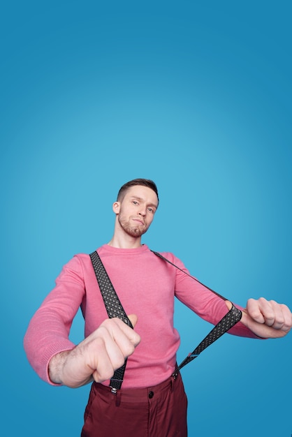 Jeune bel homme en pull et pantalon décontracté rose qui s'étend des bretelles en position debout