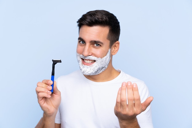 Jeune bel homme rasant sa barbe sur un mur isolé invitant à venir avec la main. Heureux que tu sois venu