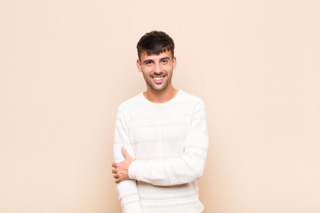 Jeune bel homme riant timidement et gaiement, avec une attitude amicale et positive mais peu sûre sur un mur chaud