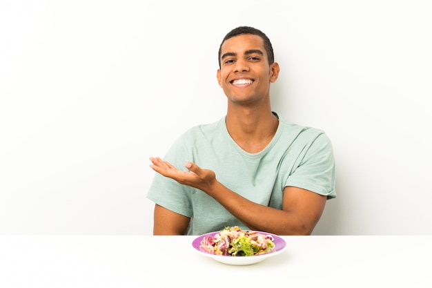 Jeune bel homme avec une salade dans une table, étendant les mains sur le côté pour avoir invité à venir