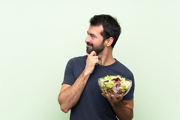 Jeune bel homme avec une salade sur le mur vert isolé, regardant sur le côté