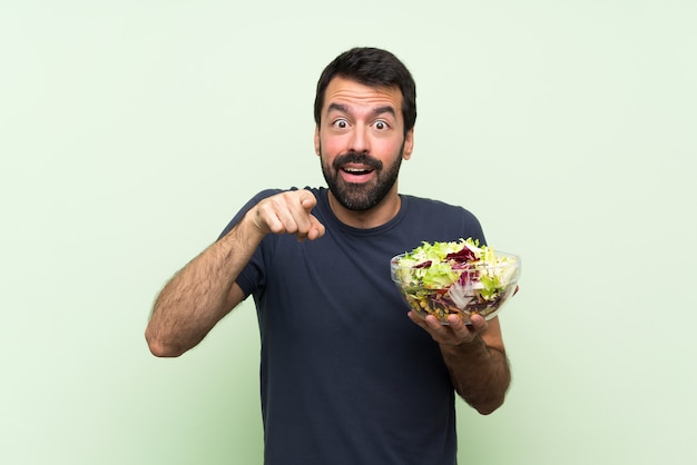 Jeune bel homme avec salade sur mur vert isolé surpris et pointant vers l'avant