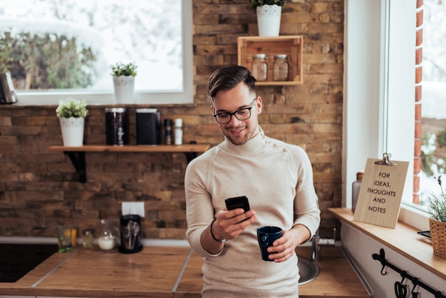 Jeune bel homme se penchant sur le comptoir de la cuisine, à l&#39;aide de téléphone et de boire du café.