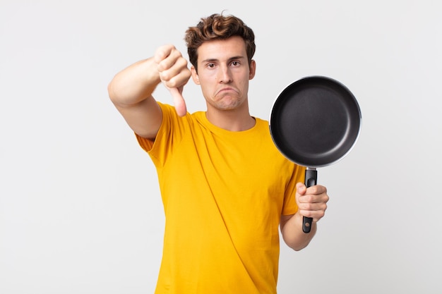 Jeune bel homme se sentant croisé, montrant les pouces vers le bas et tenant une casserole