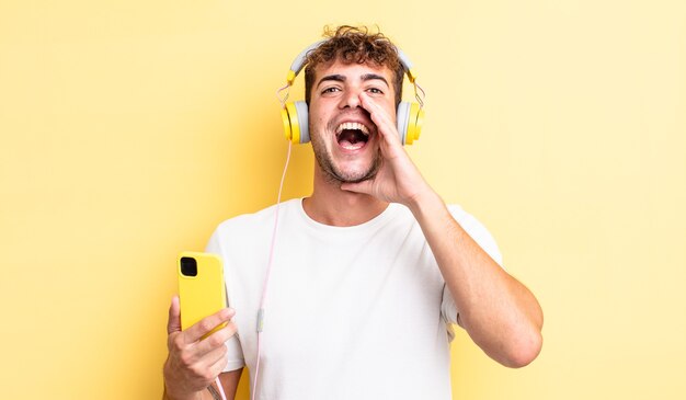 Jeune bel homme se sentant heureux, donnant un grand cri avec les mains à côté de la bouche. concept de casque et smartphone