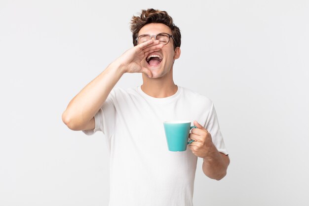 Jeune bel homme se sentant heureux, donnant un grand cri avec les mains à côté de la bouche et tenant une tasse de café