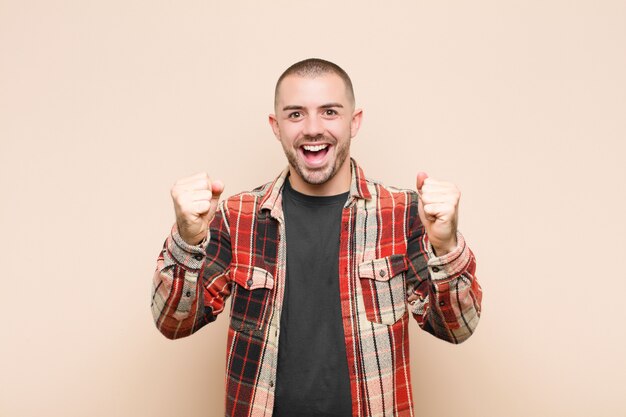 Jeune bel homme se sentant heureux, surpris et fier, criant et célébrant le succès avec un grand sourire contre le mur plat