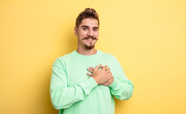 Jeune bel homme se sentant romantique, heureux et amoureux, souriant joyeusement et se tenant la main près du cœur. étrange concept de moustache