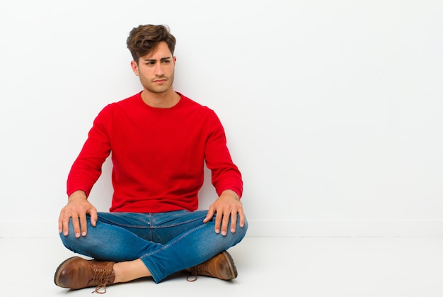 Jeune bel homme se sentant triste, contrarié ou en colère et regardant sur le côté avec une attitude négative, fronçant les sourcils en désaccord assis sur le sol