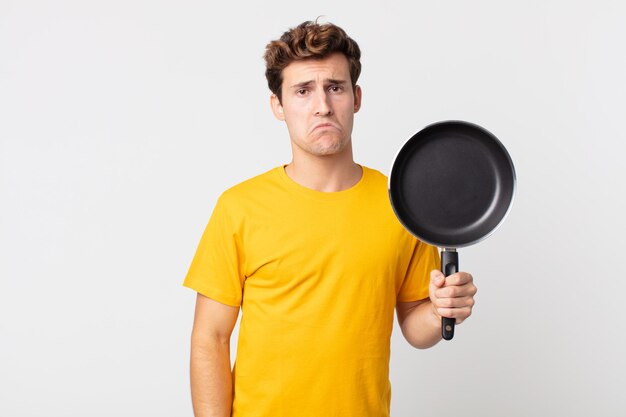 Jeune bel homme se sentant triste et geignard avec un regard malheureux et pleurant et tenant une casserole