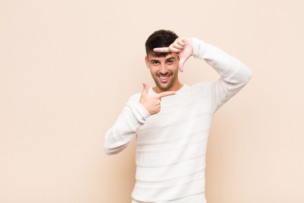 Jeune Bel Homme Se Sentir Heureux, Sympathique Et Positif, Souriant Et Faisant Un Portrait Ou Un Cadre Photo Avec Les Mains Contre Un Mur Chaud