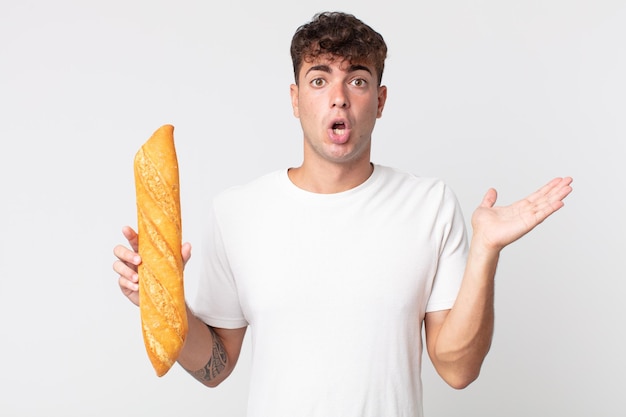 Jeune bel homme semblant surpris et choqué, avec la mâchoire tombée tenant un objet et tenant une baguette de pain