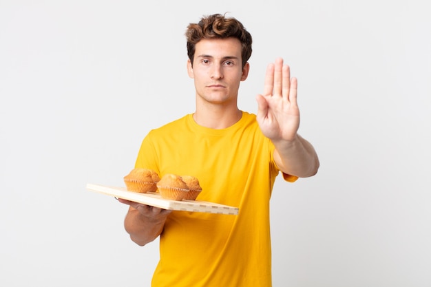 Jeune bel homme à la sérieuse montrant la paume ouverte faisant un geste d'arrêt tenant un plateau à muffins