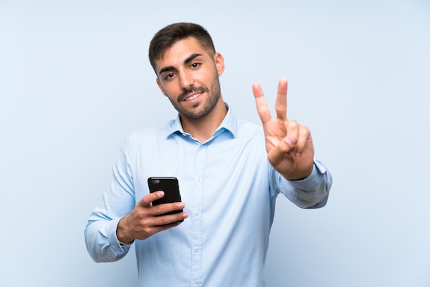 Jeune bel homme avec son téléphone portable sur un mur bleu isolé, souriant et en montrant le signe de la victoire