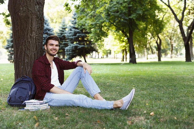 Jeune bel homme souriant assis sur l'herbe, se reposer avec des livres à proximité, rêvant de vacances, espace de copie