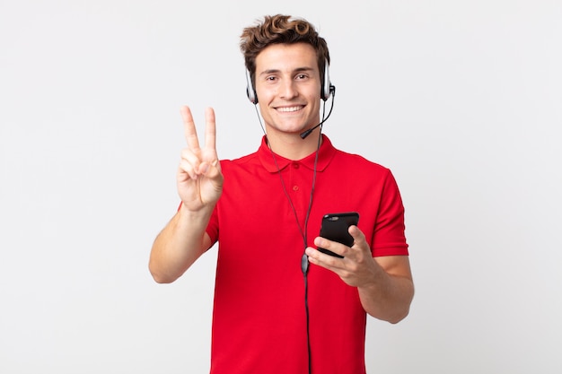 Jeune bel homme souriant et ayant l'air heureux, gesticulant la victoire ou la paix avec un smartphone et un casque