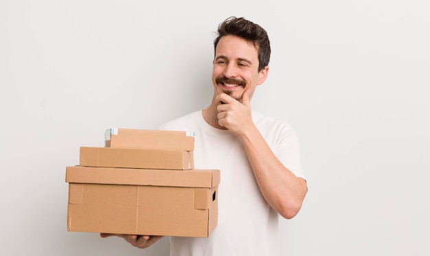 Jeune bel homme souriant avec une expression confiante heureuse avec la main sur le concept de boîtes d'expédition de menton