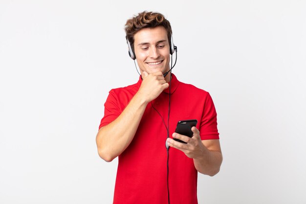 Jeune bel homme souriant avec une expression heureuse et confiante avec la main sur le menton avec un smartphone et un casque