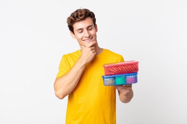 Jeune bel homme souriant avec une expression heureuse et confiante avec la main sur le menton et tenant des boîtes à lunch