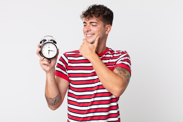 Jeune bel homme souriant avec une expression heureuse et confiante avec la main sur le menton et tenant un réveil