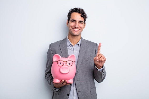 Jeune bel homme souriant fièrement et en toute confiance faisant le numéro un. notion de tirelire