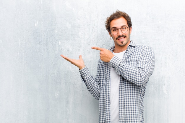 Jeune bel homme souriant gaiement et pointant pour copier l'espace sur la paume sur le côté, montrant ou annonçant un objet isolé contre un mur plat