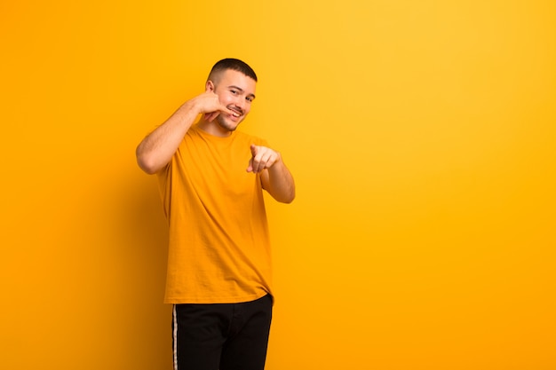 Jeune bel homme souriant gaiement tout en faisant un appel vous geste plus tard, parler au téléphone contre un mur plat