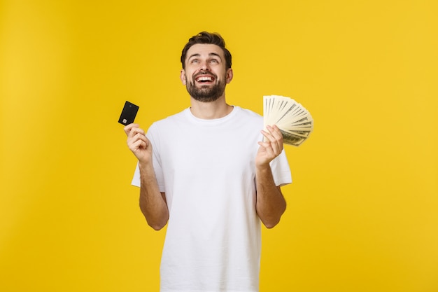 Jeune bel homme souriant heureux tenant une carte bancaire et de l'argent dans ses mains isolé sur jaune.