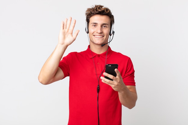 Jeune bel homme souriant joyeusement, agitant la main, vous accueillant et vous saluant avec un smartphone et un casque