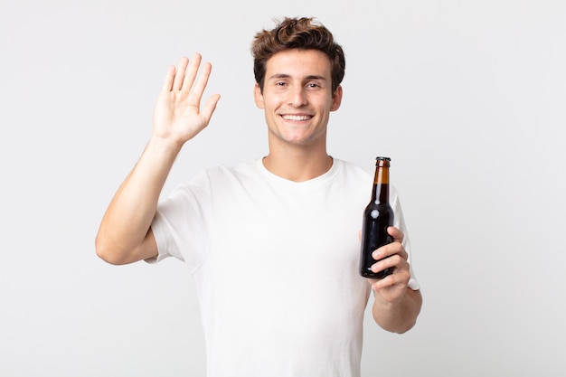 Jeune bel homme souriant joyeusement, agitant la main, vous accueillant et vous saluant et tenant une bouteille de bière