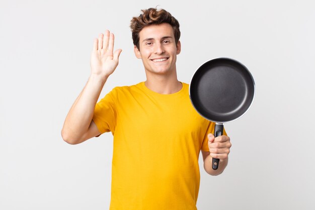 Jeune bel homme souriant joyeusement, agitant la main, vous accueillant et vous saluant et tenant une casserole