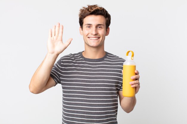 Jeune bel homme souriant joyeusement, agitant la main, vous accueillant et vous saluant et tenant un thermos à café