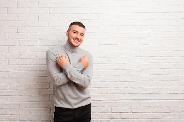 Jeune bel homme souriant joyeusement et célébrant, les poings serrés et les bras croisés, se sentant heureux et positif contre le mur plat