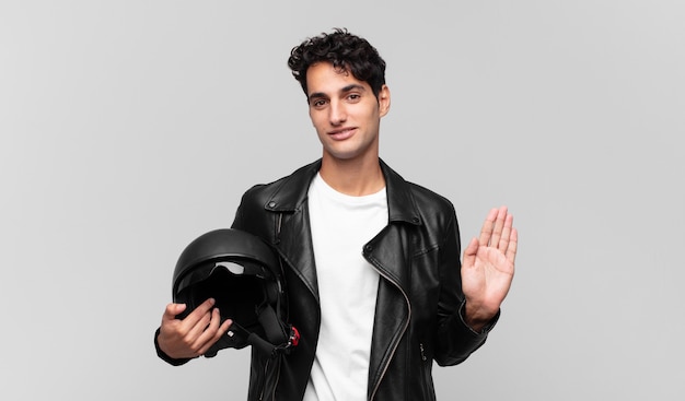 Jeune bel homme souriant joyeusement et gaiement, agitant la main, vous accueillant et vous saluant, ou vous disant au revoir. concept de pilote de moto