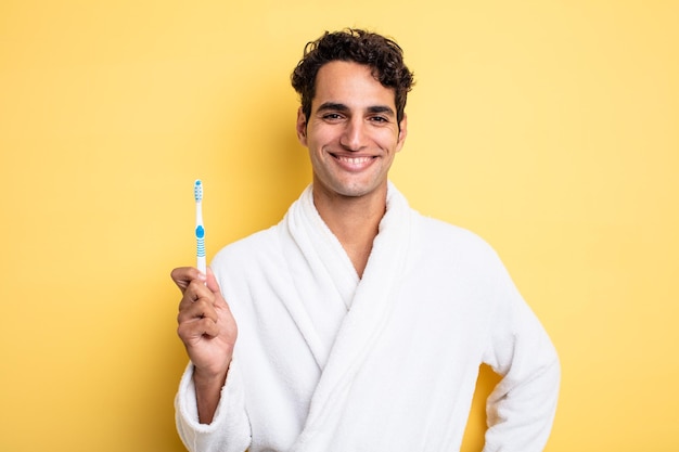 Jeune bel homme souriant joyeusement avec une main sur la hanche et un concept de peignoir et de brosse à dents confiant